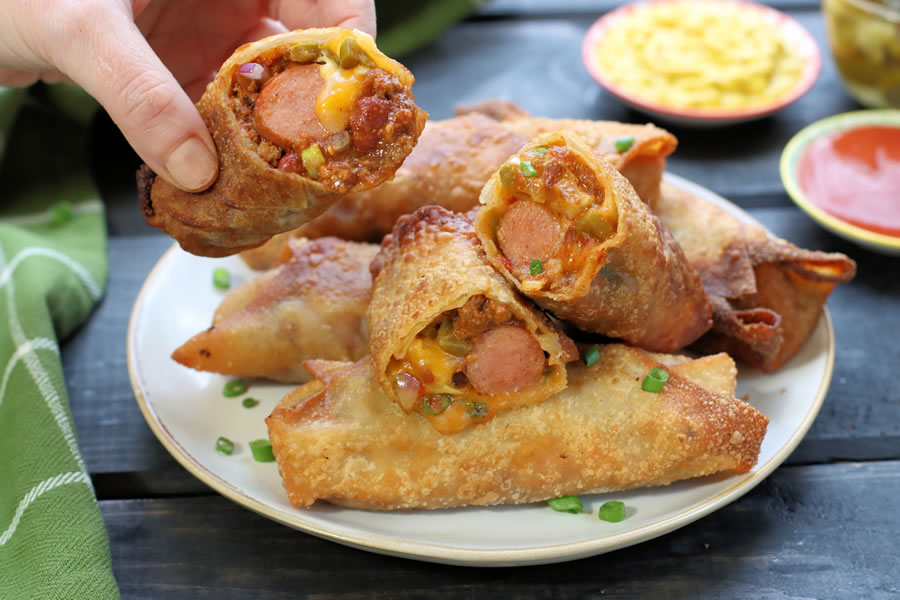 Chili Cheese Dog Egg Rolls on a plate with a hand holding one crispy fried egg roll