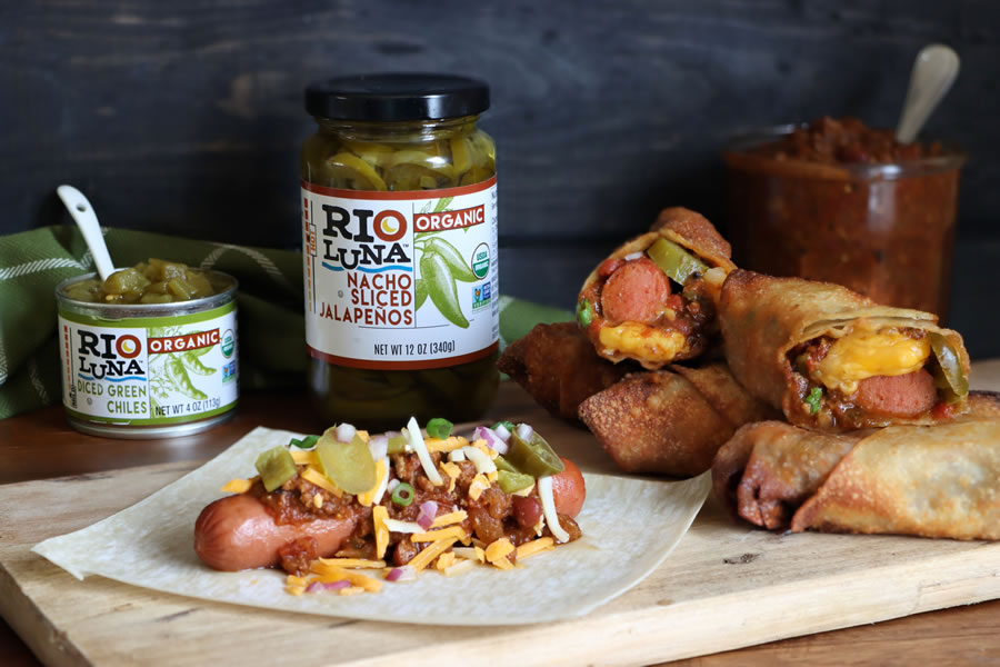 chili cheese dog being prepped with Rio Luna Nacho Sliced Jalapenos and Diced Green chilis next to finished fried egg rolls