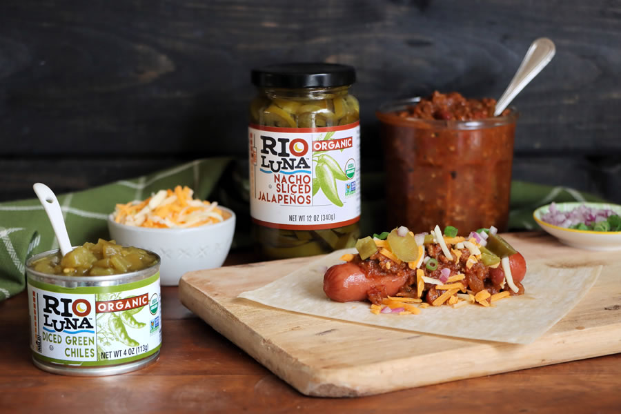chili cheese dog being assembled with Rio Luna Nacho Sliced Jalapenos and Diced Green chilis on cutting board
