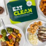 Overhead view of multiple Eat Clean meals arranged on a white marble surface. Visible dishes include pulled pork, sausage with vegetables, surf and turf with potatoes, meatballs, chicken nuggets with mac and cheese, and mini churro donuts. The plates feature different protein options, sides, and colorful vegetables, demonstrating the variety available in their meal plans.
