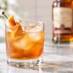 close-up of a SoCo amber-colored cocktail served over large ice cubes in a clear rocks glass on a pale marble countertop. The chilled glass is covered with natural condensation and garnished with a curled orange peel on a decorative cocktail pick. A Southern Comfort bottle sits softly blurred in the background, while natural daylight and a softly out-of-focus plant create a bright, warm kitchen-bar setting.