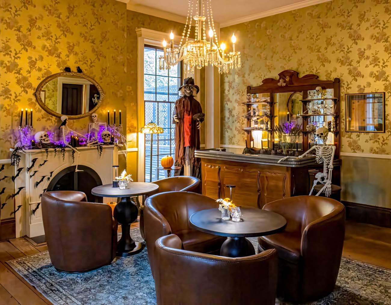 Elegant Manor House lounge transformed for Halloween with antique wooden bar, skeleton bartender prop, life-size witch figure in Victorian dress, crystal chandelier, gold wallpaper, fireplace decorated with black paper bats, purple LED lights, black candles on mantel, leather club chairs around small tables, oriental rug on hardwood floors