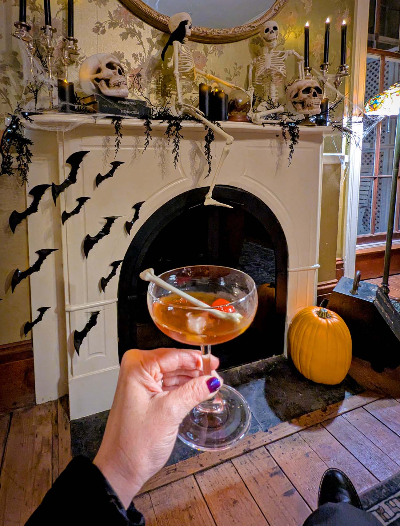 Elegant fireplace mantel decorated with skeleton figures, black candles, paper bats on walls, bourbon cocktail with bone stirrer held in foreground, pumpkin on floor