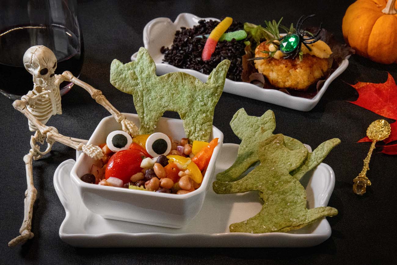 Halloween-themed bean salads in white square dishes with green cat-shaped tortilla chips and googly eyes as garnish, plastic skeleton decoration, gold spoon on black tablecloth