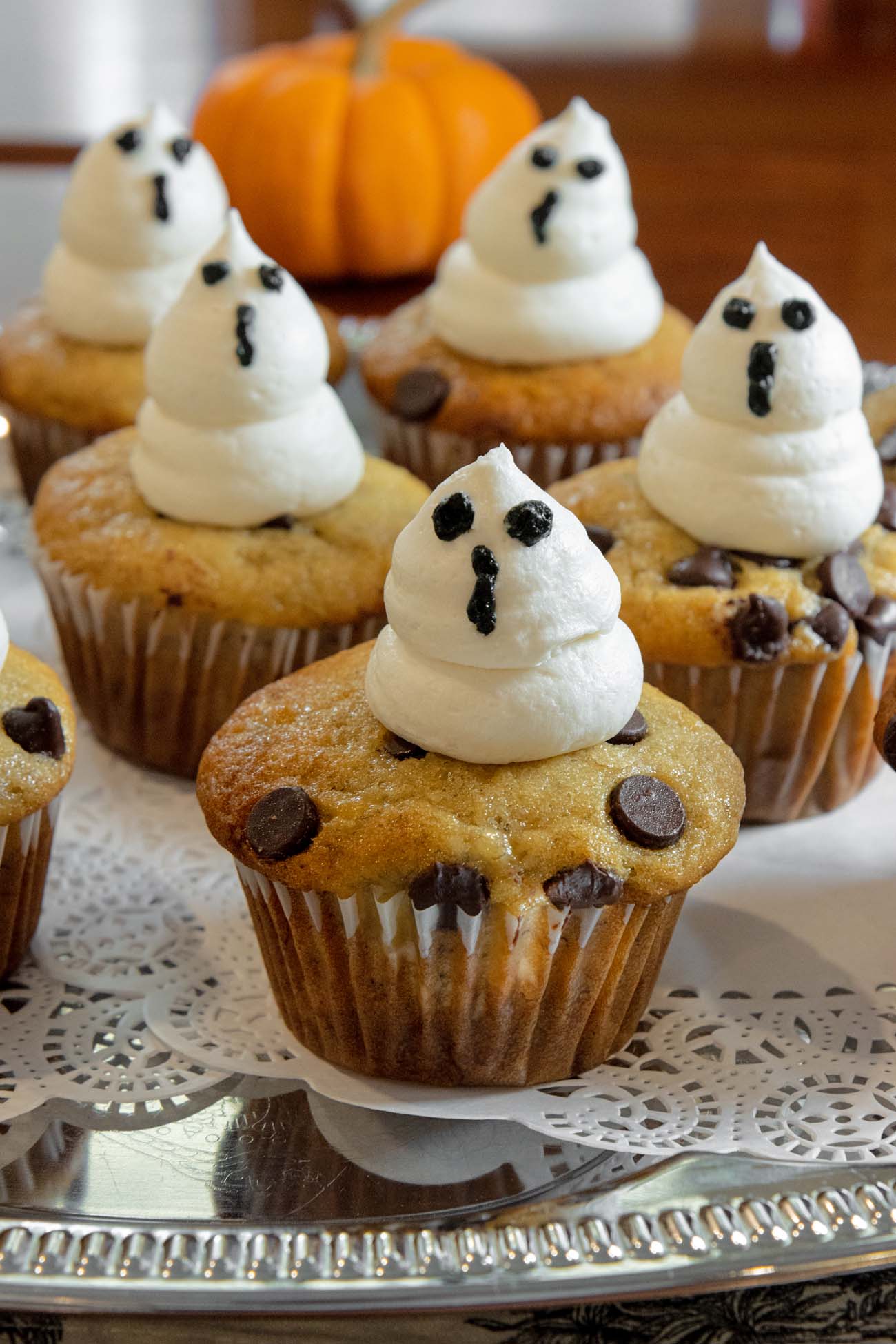 Chocolate chip muffins decorated with white frosting ghosts with chocolate chip eyes and mouths, arranged on silver serving tray with pumpkin in background