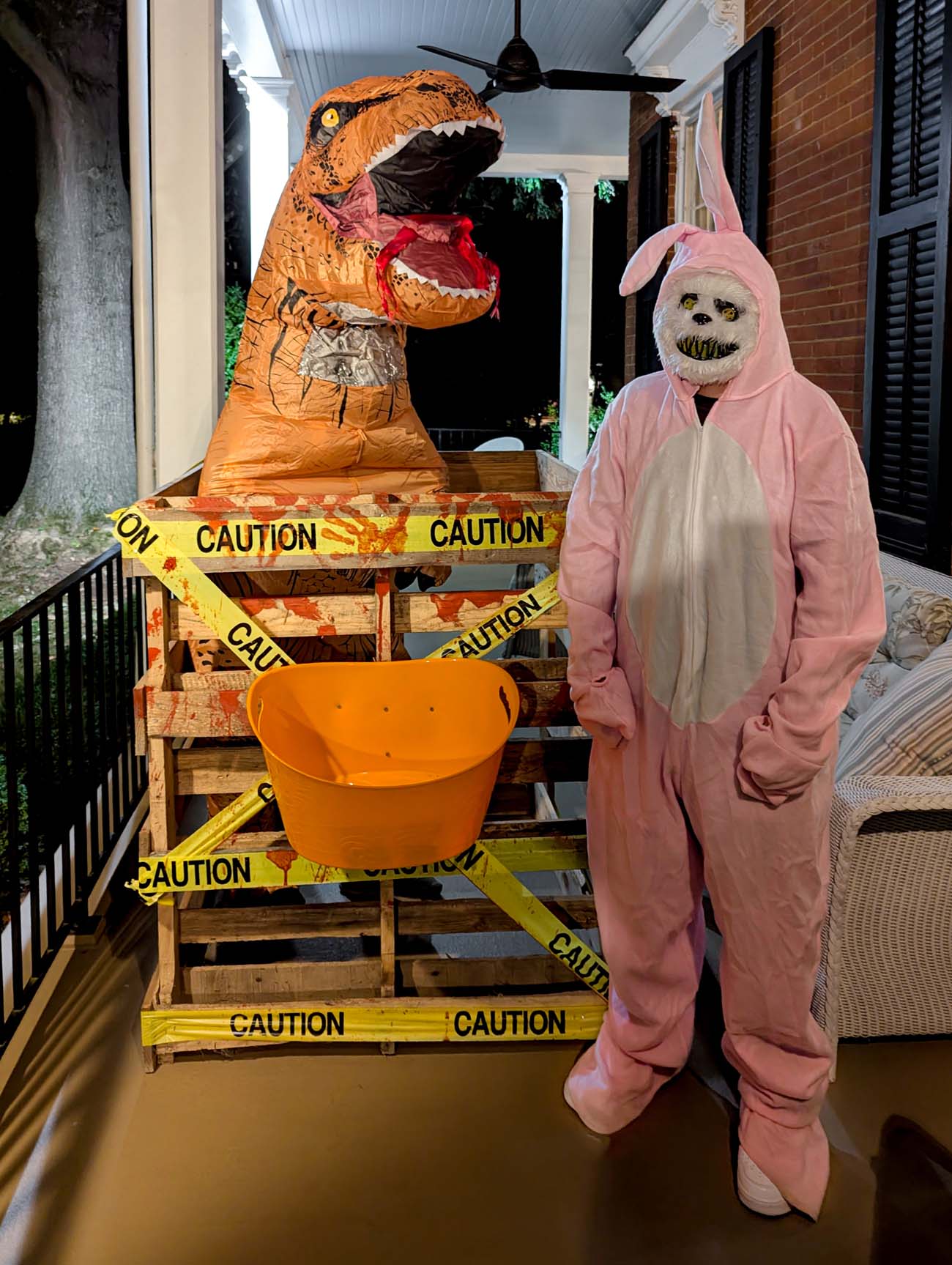 Inflatable T-Rex costume and person in pink bunny onesie with scary face standing on porch with orange candy bucket and caution tape decoration