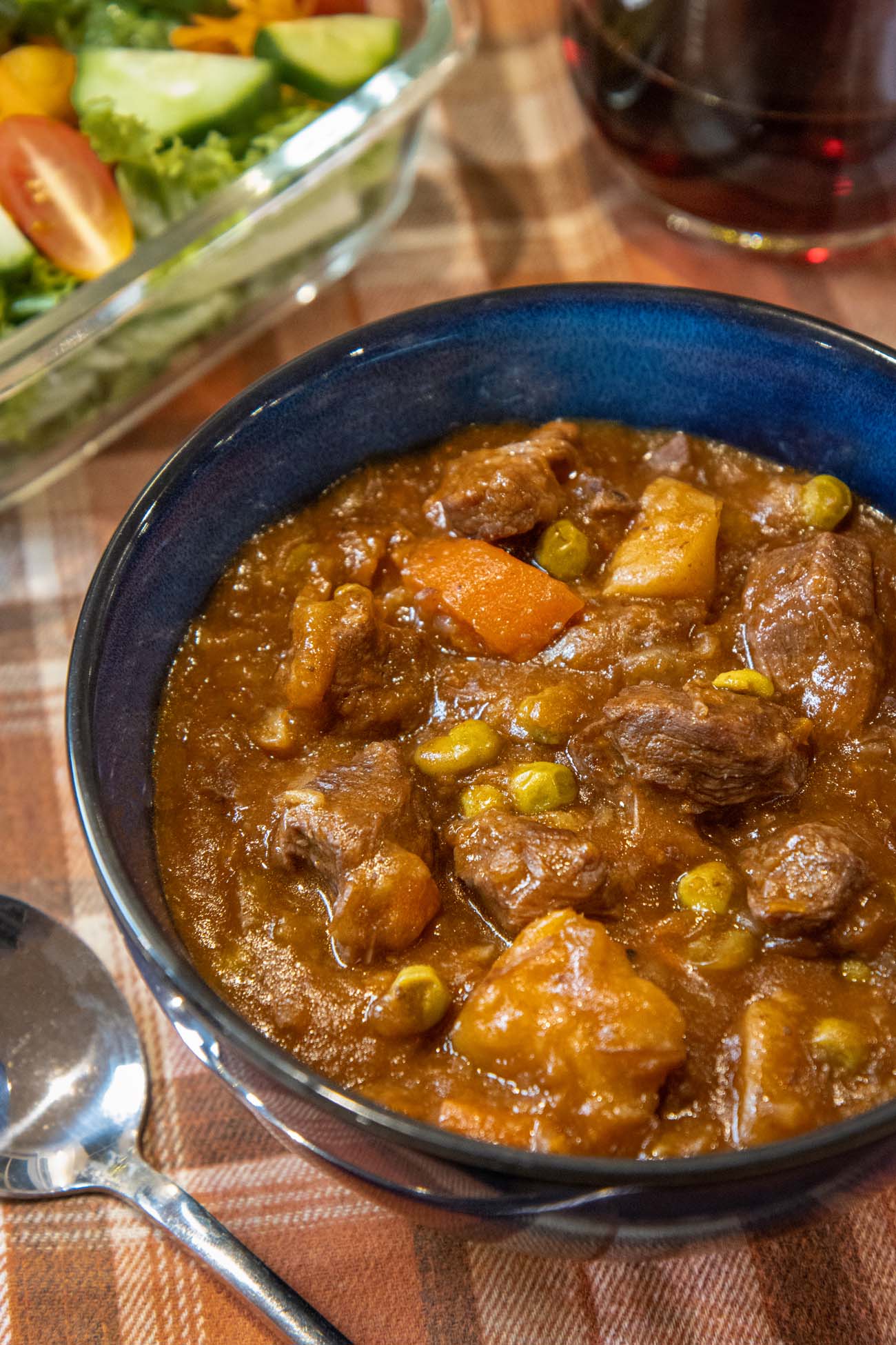 Bowl of rich beef stew with chunks of meat, carrots, potatoes and peas in thick gravy, served with side salad and red wine on woven placemat