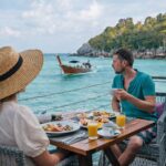 a man on woman dining dockside in Thailand as a wooden boat drifts by