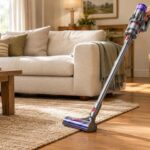 A cordless vacuum with a purple canister and floor head cleans a woven rug beside a beige sofa in a bright, sunlit living room.